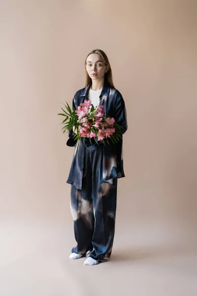 Piena lunghezza della donna in abito stile tie-dye in piedi con bouquet floreale su sfondo beige — Foto stock
