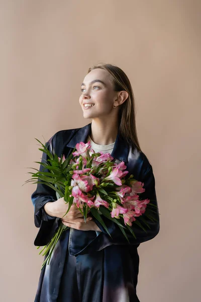 Счастливая и модная женщина со свежим цветочным букетом, смотрящая вдаль на бежевый — стоковое фото