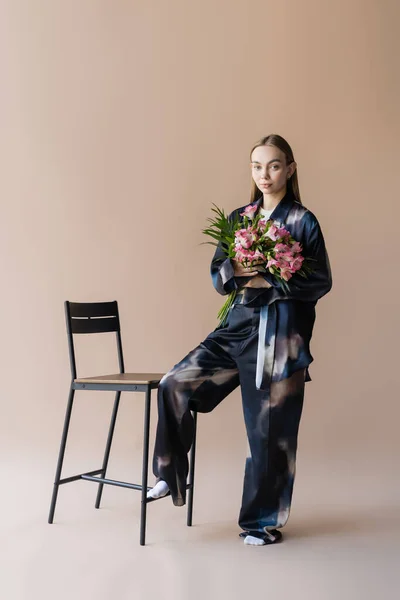 Femme à la mode avec des fleurs alstroemeria regardant la caméra près de la chaise sur fond beige — Photo de stock