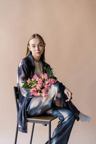 Young woman in gradient clothes sitting on chair with alstroemeria flowers isolated on beige — Stockfoto