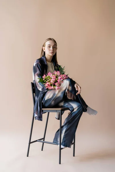 Full length of stylish woman with alstroemeria bouquet posing on chair on beige background — Foto stock