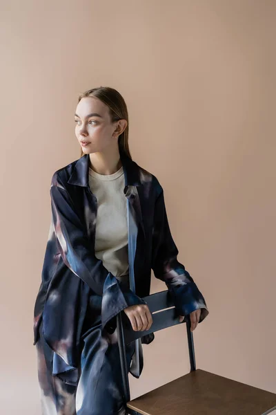 Young woman in tie-dye shirt looking away near chair isolated on beige — Photo de stock