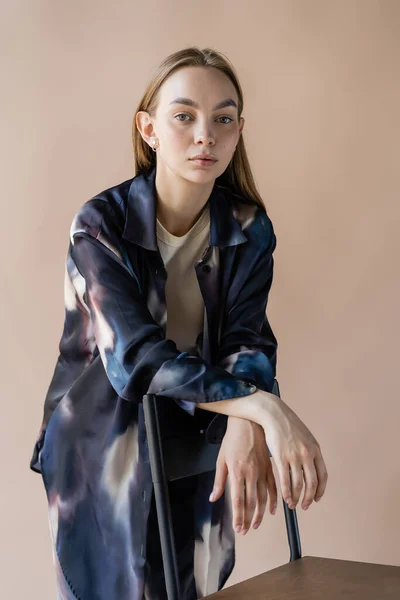 Pretty woman in trendy oversize shirt posing near chair isolated on beige — Stock Photo