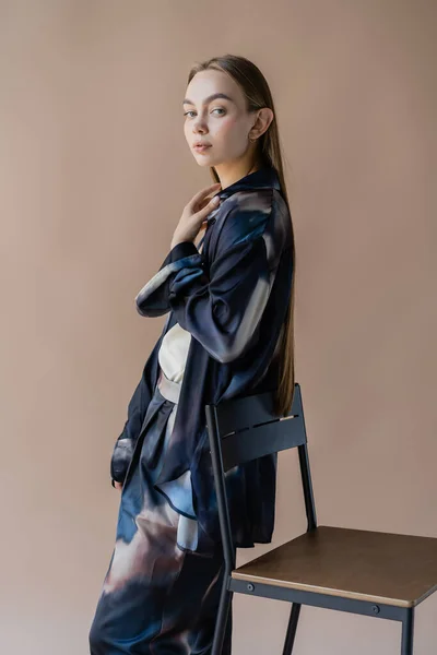 Woman in trendy oversize clothing looking at camera near chair isolated on beige - foto de stock