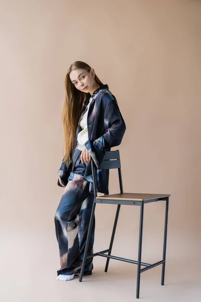 Pleine longueur de femme aux cheveux longs élégant posant près de la chaise sur fond beige — Photo de stock