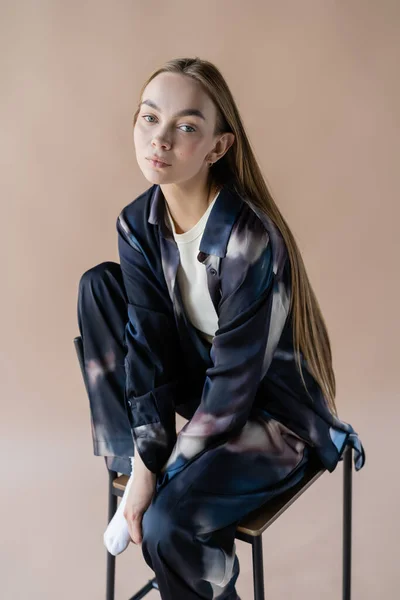 Jeune femme aux cheveux longs dans des vêtements de style tie-dye posant sur chaise isolée sur beige — Photo de stock