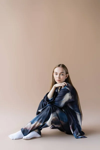 Young woman in gradient clothing sitting with hand near chin on beige background — Photo de stock