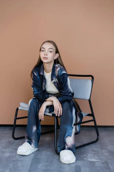 Full length of woman in trendy clothes and fluffy slippers sitting on modern chair on beige — Stockfoto