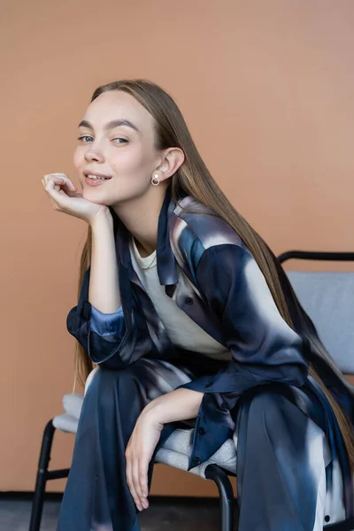 Mujer positiva en ropa de estilo tie-dye sentada con la mano cerca de la barbilla sobre fondo beige - foto de stock