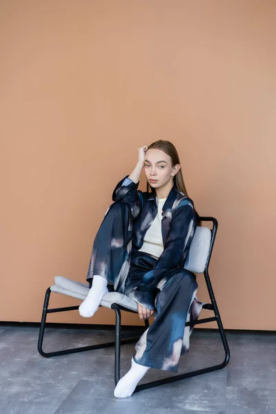 Pleine longueur de la femme en vêtements oversize posant sur chaise moderne sur fond beige — Photo de stock