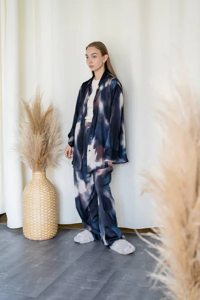 Full length of woman in oversize suit and fluffy slippers near wicker vase with spikelets and white drapery - foto de stock