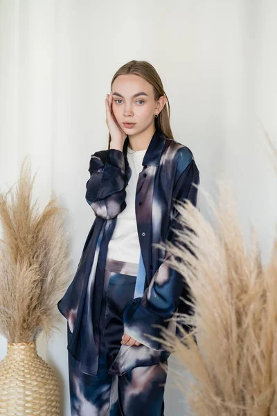 Trendy woman in tie-dye style clothes touching face near dried spikelets on white background — Photo de stock