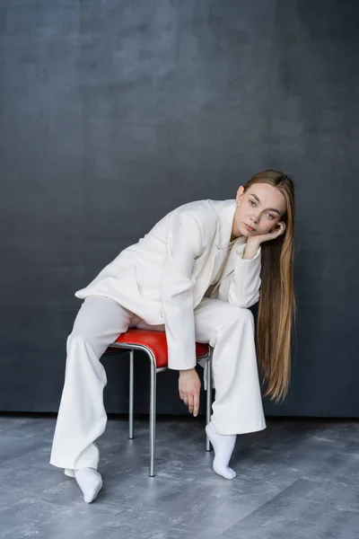 Volle Länge der stilvollen Frau in weißem Anzug und Socken sitzt auf Stuhl auf schwarzem Hintergrund — Stockfoto