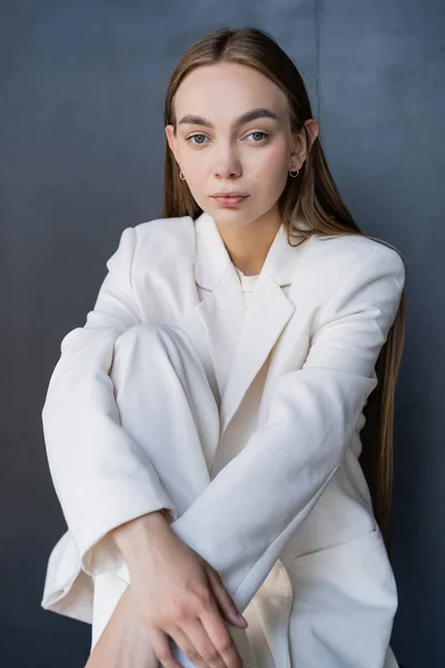 Young woman in white trendy clothes looking at camera while sitting on black background - foto de stock