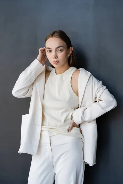 Femme en costume blanc et débardeur debout avec la main sur la hanche et la fixation des cheveux sur fond noir — Photo de stock