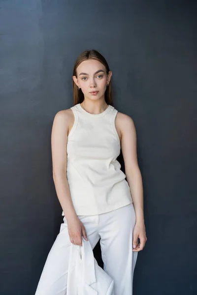 Slim woman in white tank top holding blazer and looking at camera on black background — Stock Photo