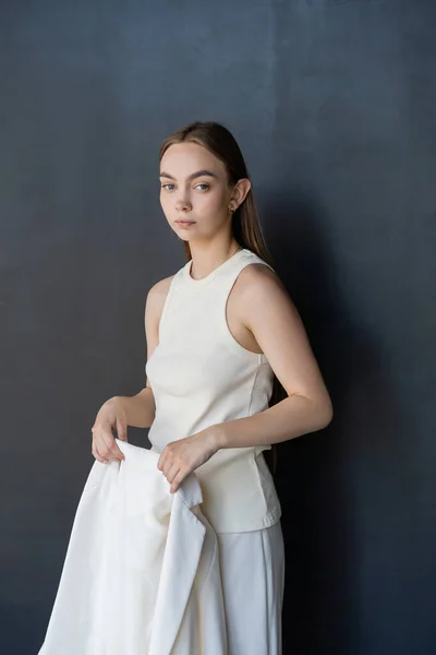 Pretty woman in white tank top holding blazer and looking at camera on black background — Fotografia de Stock