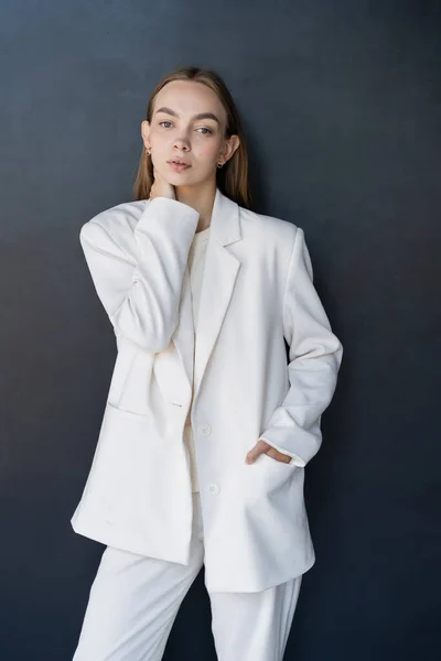 Stylish woman in white blazer touching neck while posing with hand in pocket on black background — Photo de stock