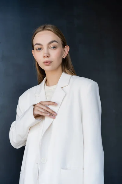 Fashionable woman touching white blazer and looking at camera on black background — Fotografia de Stock