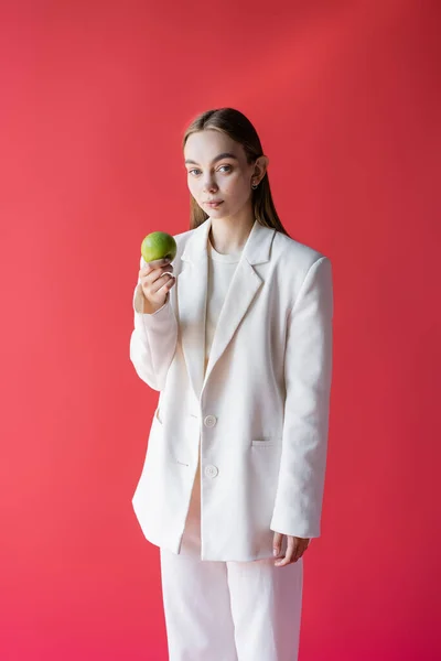 Pretty woman in white blazer holding fresh apple and looking at camera isolated on crimson - foto de stock