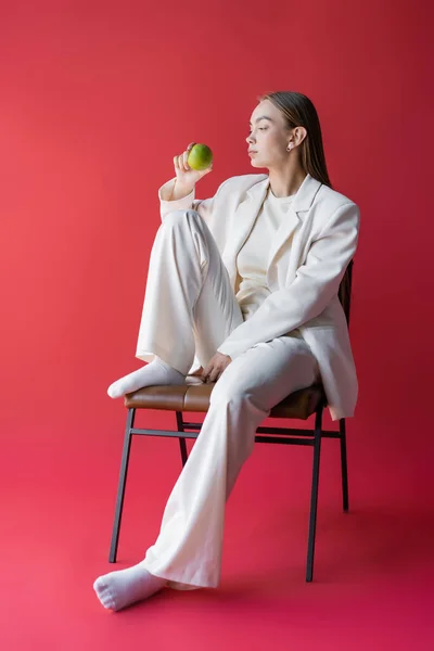 Full length of woman in white suit and socks sitting on chair with apple on pink background — Foto stock