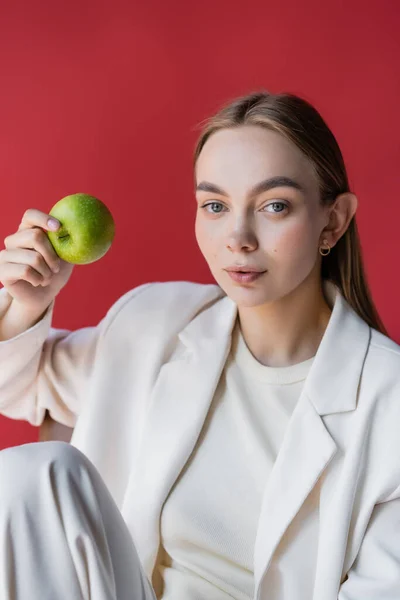Красивая и модная женщина с сочным яблоком, смотрящая на камеру, изолированную от малинового — стоковое фото