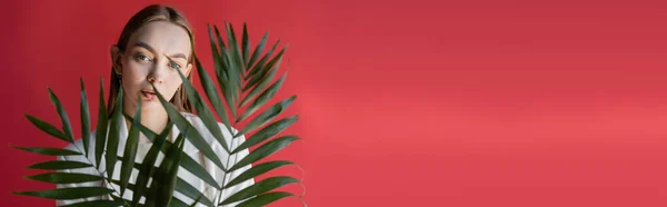 Charmante femme en vêtements blancs regardant la caméra à travers des feuilles tropicales isolées sur rose, bannière — Photo de stock