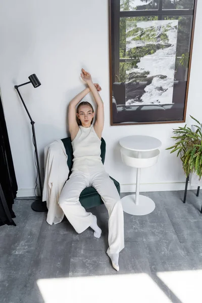 Full length of woman in white clothes sitting in armchair with raised hands near coffee table — Foto stock