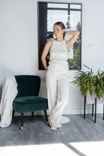 Full length of woman in white posing near black armchair and looking away — Foto stock