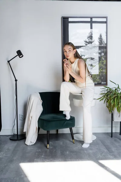 Full length of woman in white clothes looking at camera near black armchair in modern room - foto de stock