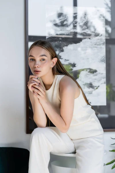 Dreamy woman in white clothes sitting on coffee table with hands near chin — Stockfoto