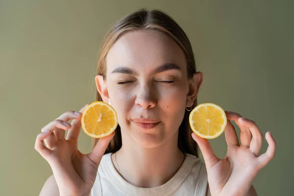 Grimacing woman with closed eyes holding halves of sour lemon isolated on green — Stockfoto