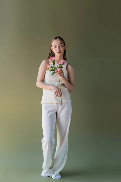 Longueur totale de la femme en pantalon blanc et débardeur debout avec des fleurs roses sur fond vert — Photo de stock
