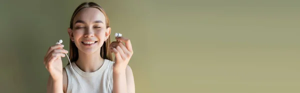 Mulher alegre e bonita com olhos fechados segurando fones de ouvido com fio isolado no verde, banner — Fotografia de Stock