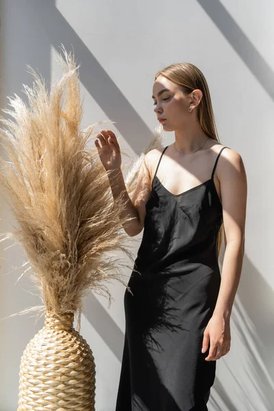 Young woman in black strap dress touching spikelets in wicker vase on white background with shadows — Photo de stock
