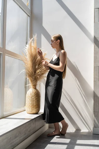 Piena lunghezza della donna a piedi nudi in abito nero vicino alla finestra e vaso di vimini con spighette — Foto stock