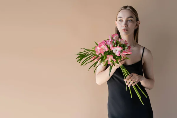 Jeune femme en robe à bretelles noire tenant bouquet de fleurs roses isolées sur beige — Photo de stock
