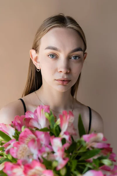 Young woman with perfect skin looking at camera near alstroemeria flowers isolated on beige — Stock Photo