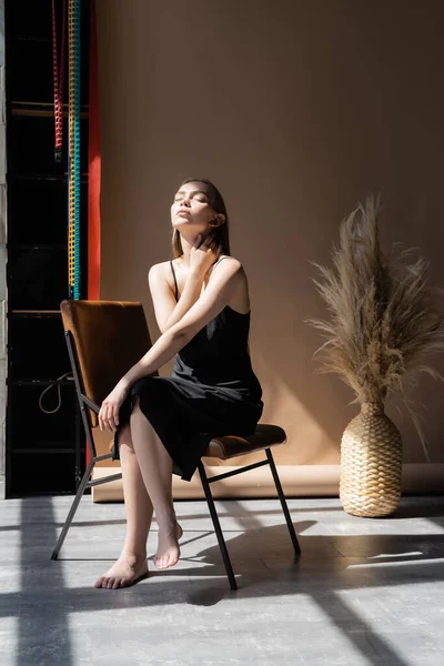 Full length of barefoot woman in black strap dress sitting with closed eyes on beige background - foto de stock