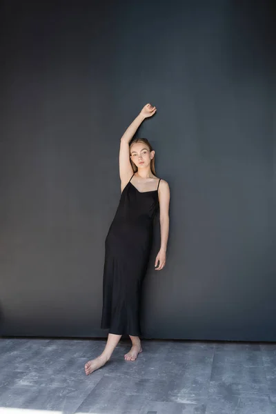 Full length of barefoot woman in black strap dress posing with raised hand near dark wall — Fotografia de Stock