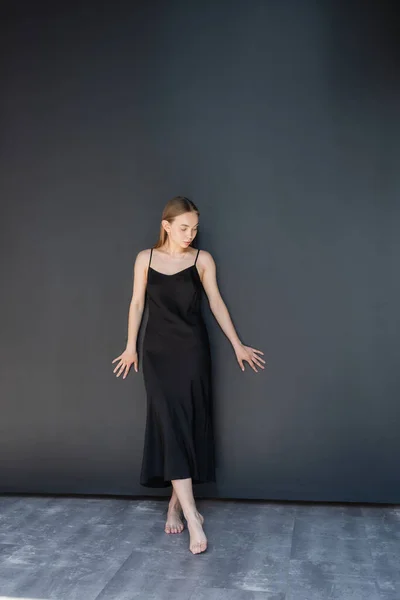 Piena lunghezza della giovane donna a piedi nudi in abito cinturino in piedi vicino alla parete nera — Foto stock