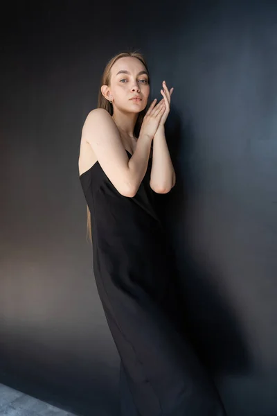 Slim and seductive woman in black strap dress looking at camera near dark wall - foto de stock