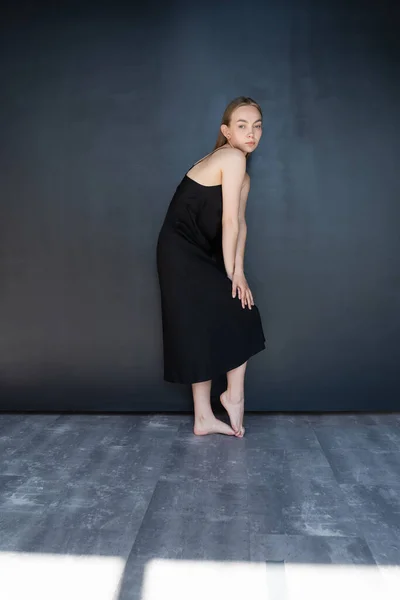 Full length of slim barefoot woman in black strap dress near dark wall — Stock Photo