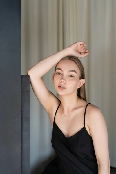 Seductive woman in black strap dress posing with hand above head near beige drapery — Stock Photo