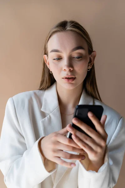 Pretty woman in white blazer using smartphone isolated on beige — Photo de stock
