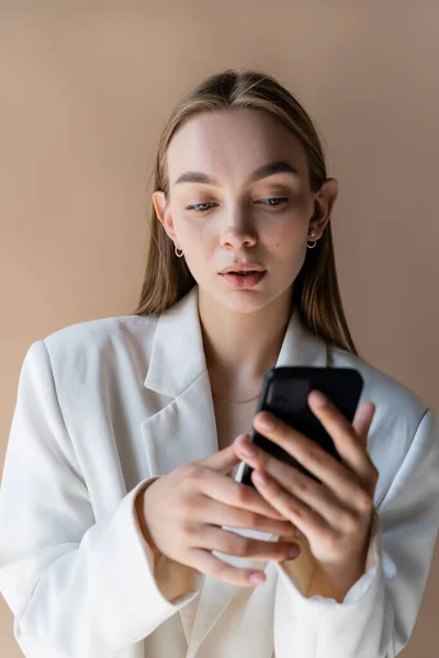 Pretty woman in white blazer using smartphone isolated on beige — Stock Photo