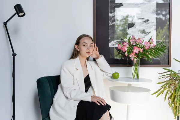 Stylish woman in white blazer sitting near apple and alstroemeria flowers on coffee table — Photo de stock