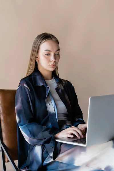 Junge Frau in stylischem Oversize-Hemd sitzt und tippt auf Laptop isoliert auf beige — Stockfoto