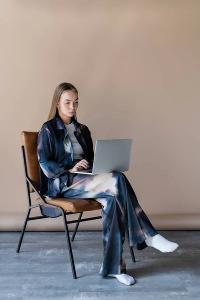 Woman in trendy oversize clothes sitting on chair and typing on laptop on beige background — Stockfoto