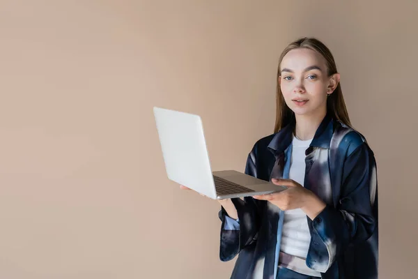 Frau in stylischem Oversize-Shirt blickt in die Kamera, während sie ihren Laptop isoliert auf beige hält — Stockfoto